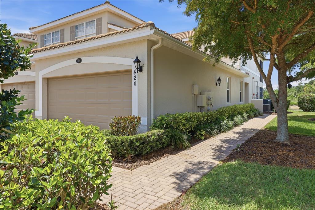 A tan two-story townhouse with a garage, a paved walkway, and landscaping under a blue sky — 424 WINDING BROOK LANE UNIT 104, BRADENTON, FL