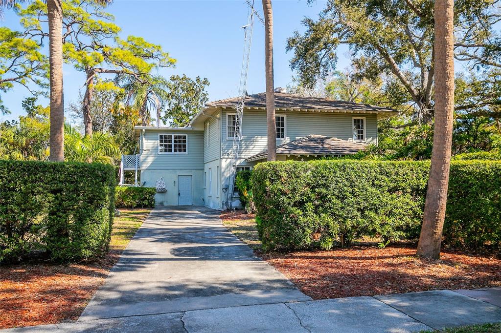 A two-story house with light green siding and a concrete driveway sits behind tall hedges — 5013 BAY SHORE ROAD, SARASOTA, FL