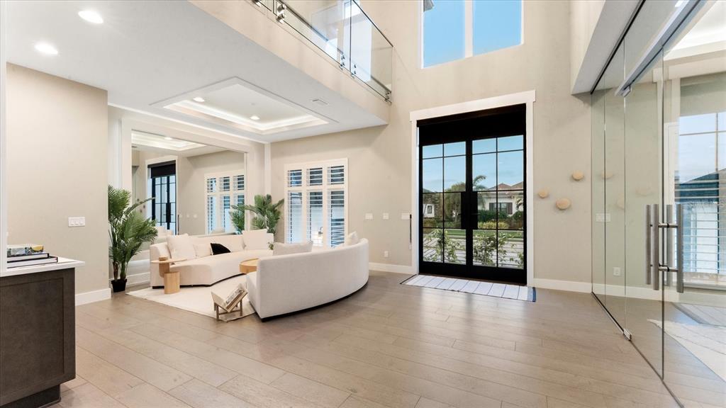 A modern, high-ceilinged living room features white curved sofas, light wood flooring, and glass walls — 5708 INSPIRATION TERRACE, BRADENTON, FL