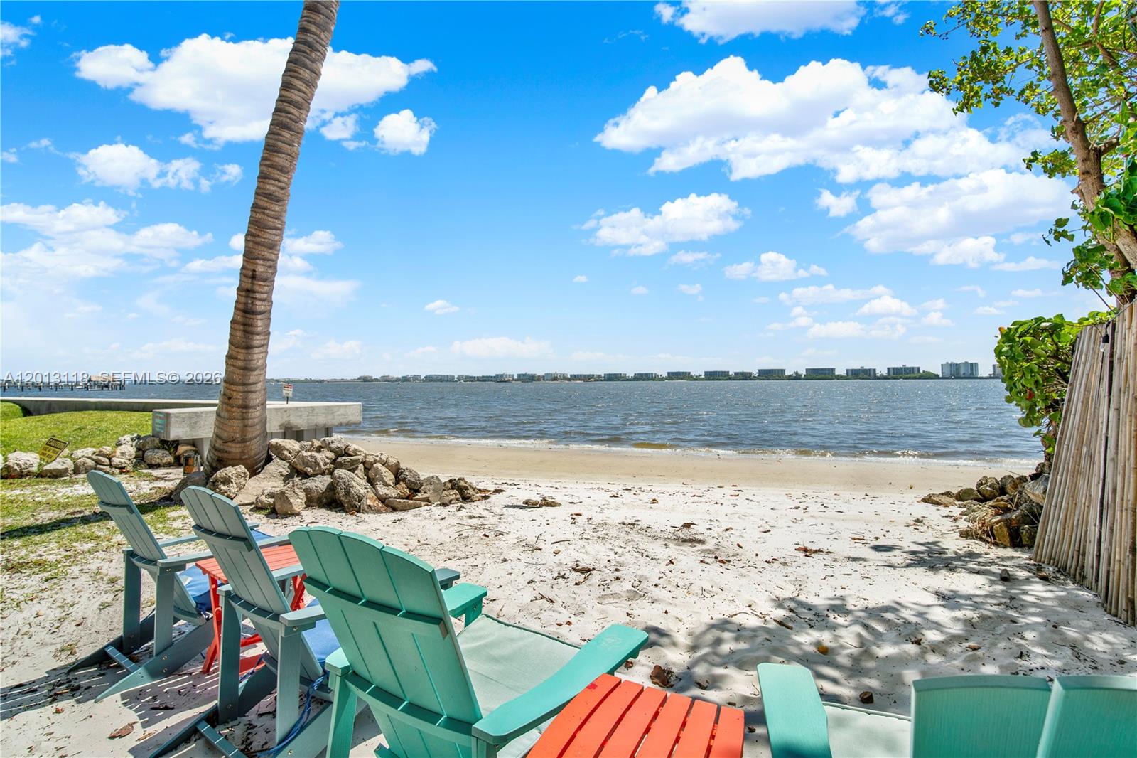 Turquoise Adirondack chairs sit on a sandy beach overlooking a bay with distant buildings — 1516 S Lakeside Dr # 105, Lake Worth FL 33460, LAKE WORTH, FL