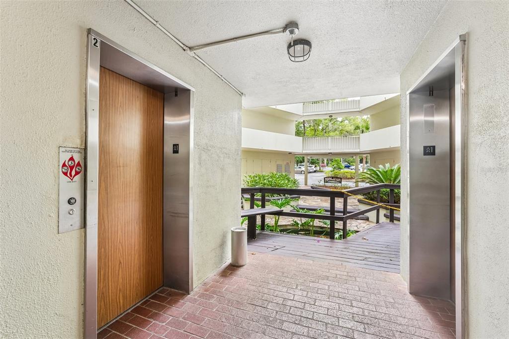 Two elevators face each other in a covered outdoor walkway with brick flooring — 1717 NW 23RD AVENUE UNIT 4E, GAINESVILLE, FL