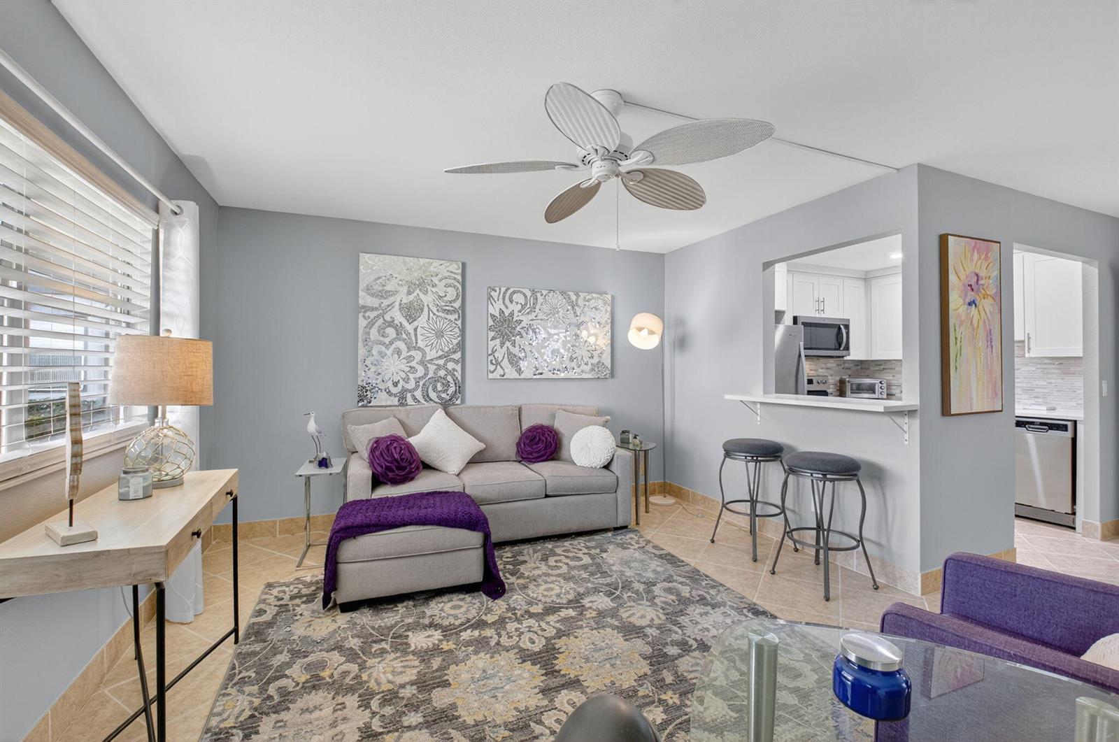 A living room features a grey sofa, purple accents, a ceiling fan, and a kitchen pass-through — 499 Monaco K # K, Delray Beach FL 33446, DELRAY BEACH, FL