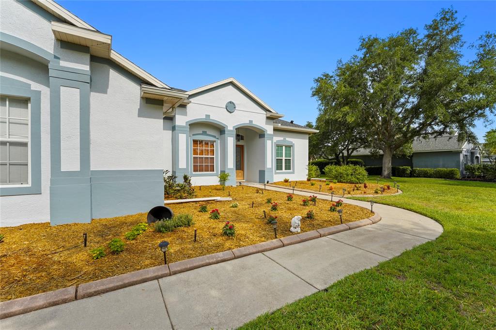 A single-story house exterior features a concrete walkway, landscaped mulch beds, and a large tree — 8018 SNOWY EGRET PLACE, BRADENTON, FL