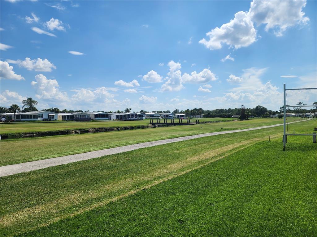 A grassy field features a paved path, a small bridge, and distant residential homes — 74 STRAPHMORE DRIVE, HAINES CITY, FL