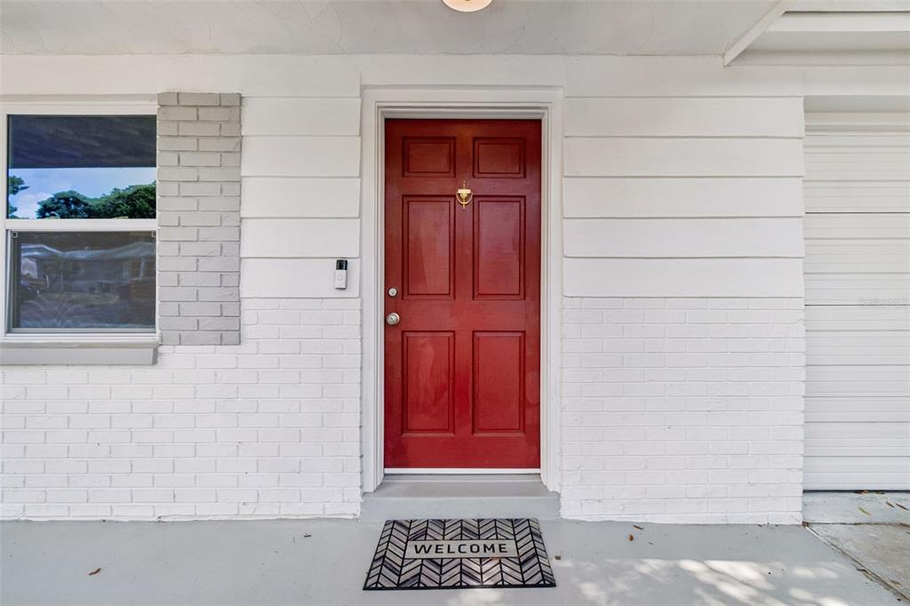 A red front door with a welcome mat is centered on a white brick exterior — 5046 FARLEY DRIVE, HOLIDAY, FL