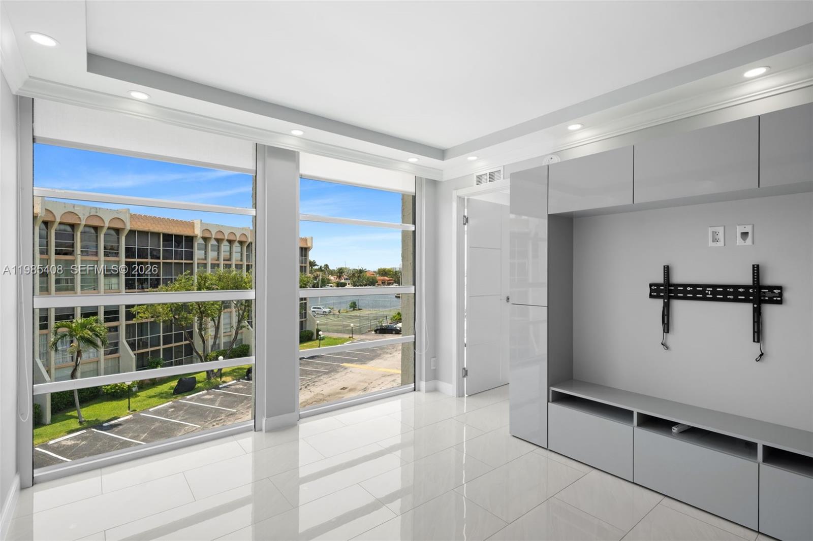 A modern room features white tile floors, a grey built-in media unit, and large windows — 701 Three Islands Blvd # 411, Hallandale Beach FL 33009, HALLANDALE BEACH, FL