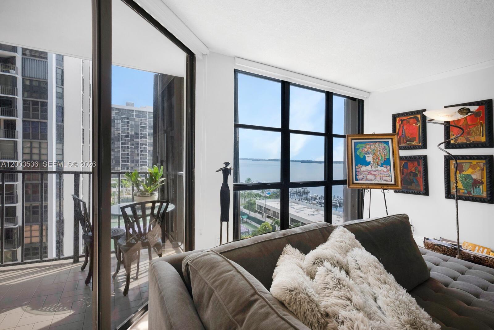 A living room features a gray sofa, floor-to-ceiling windows, and a balcony overlooking the water — 1925 Brickell Ave # 1003, Miami FL 33129, MIAMI, FL