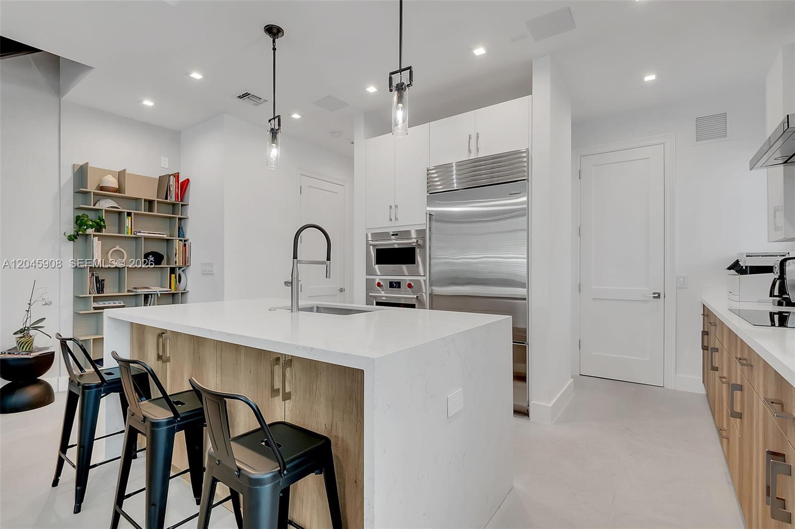 A modern kitchen features a white quartz island, wood-toned cabinetry, and black metal bar stools — 3406 Ponce De Leon Blvd, Coral Gables FL 33134, CORAL GABLES, FL