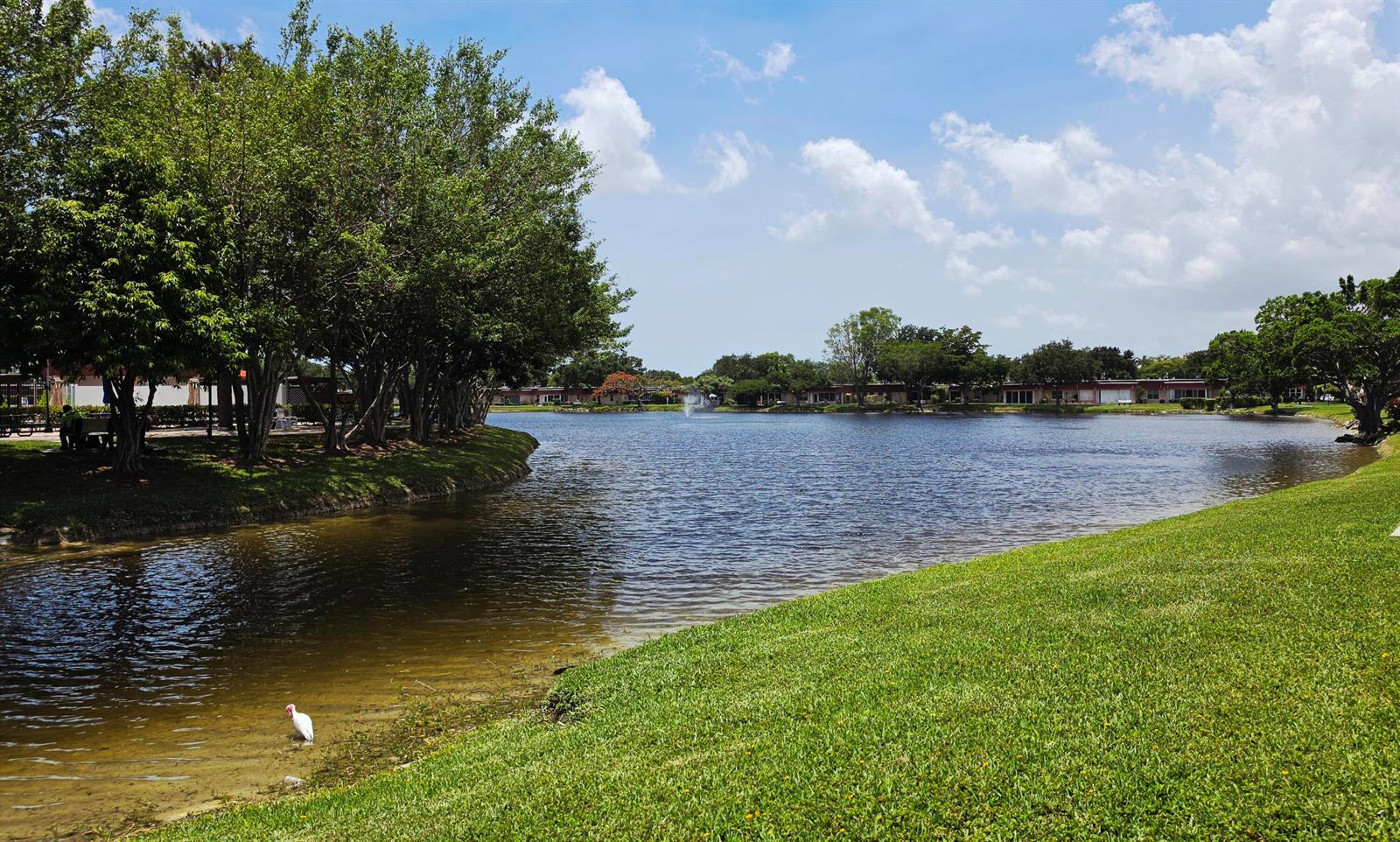 A pond with a fountain is surrounded by green grass and trees near residential buildings — 713 Nantucket Circle # B, Lake Worth FL 33467, LAKE WORTH, FL