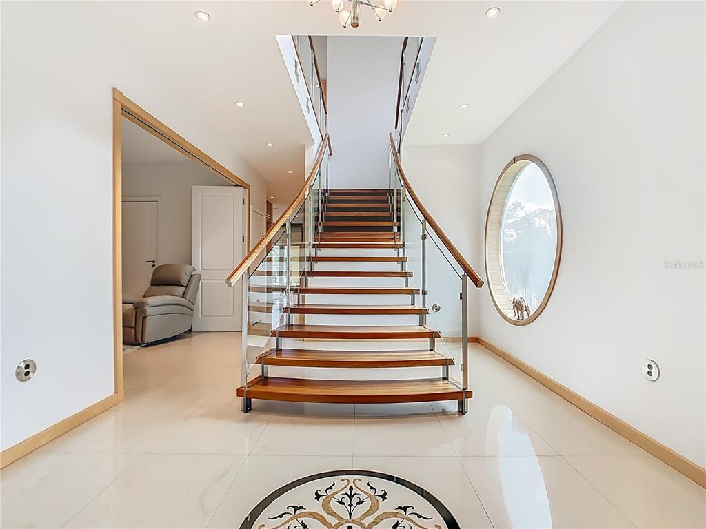 A modern foyer features a floating wooden staircase with glass railings and a circular window — 917 DESERT MOUNTAIN COURT, REUNION, FL
