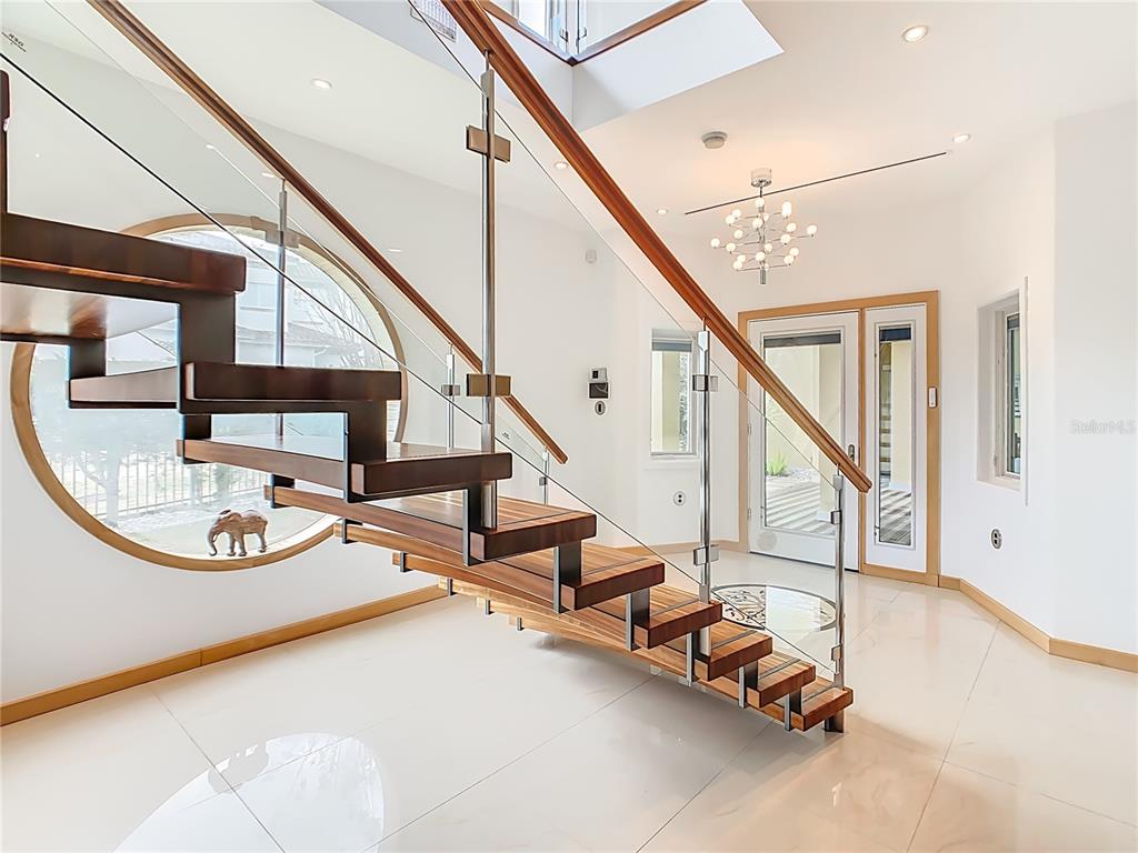 A modern foyer features a floating wooden staircase with glass railings and a circular window — 917 DESERT MOUNTAIN COURT, REUNION, FL