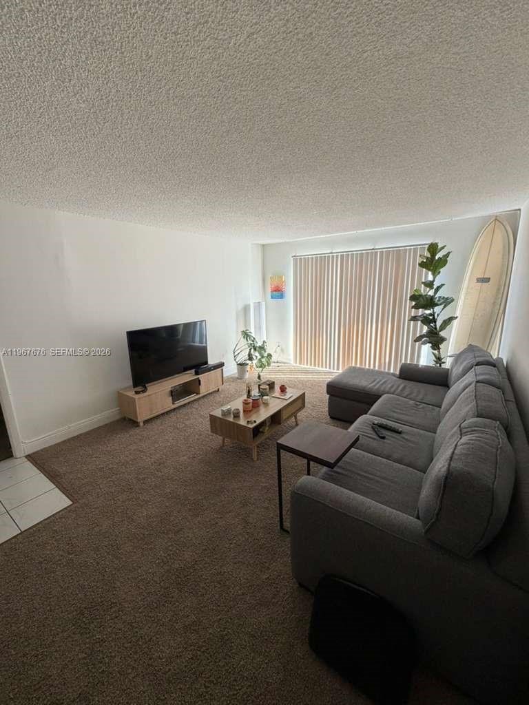 A living room with carpeted floors, a gray sectional sofa, a television, and a surfboard — 488 NW 165th St Rd # 614, Miami FL 33169, MIAMI, FL