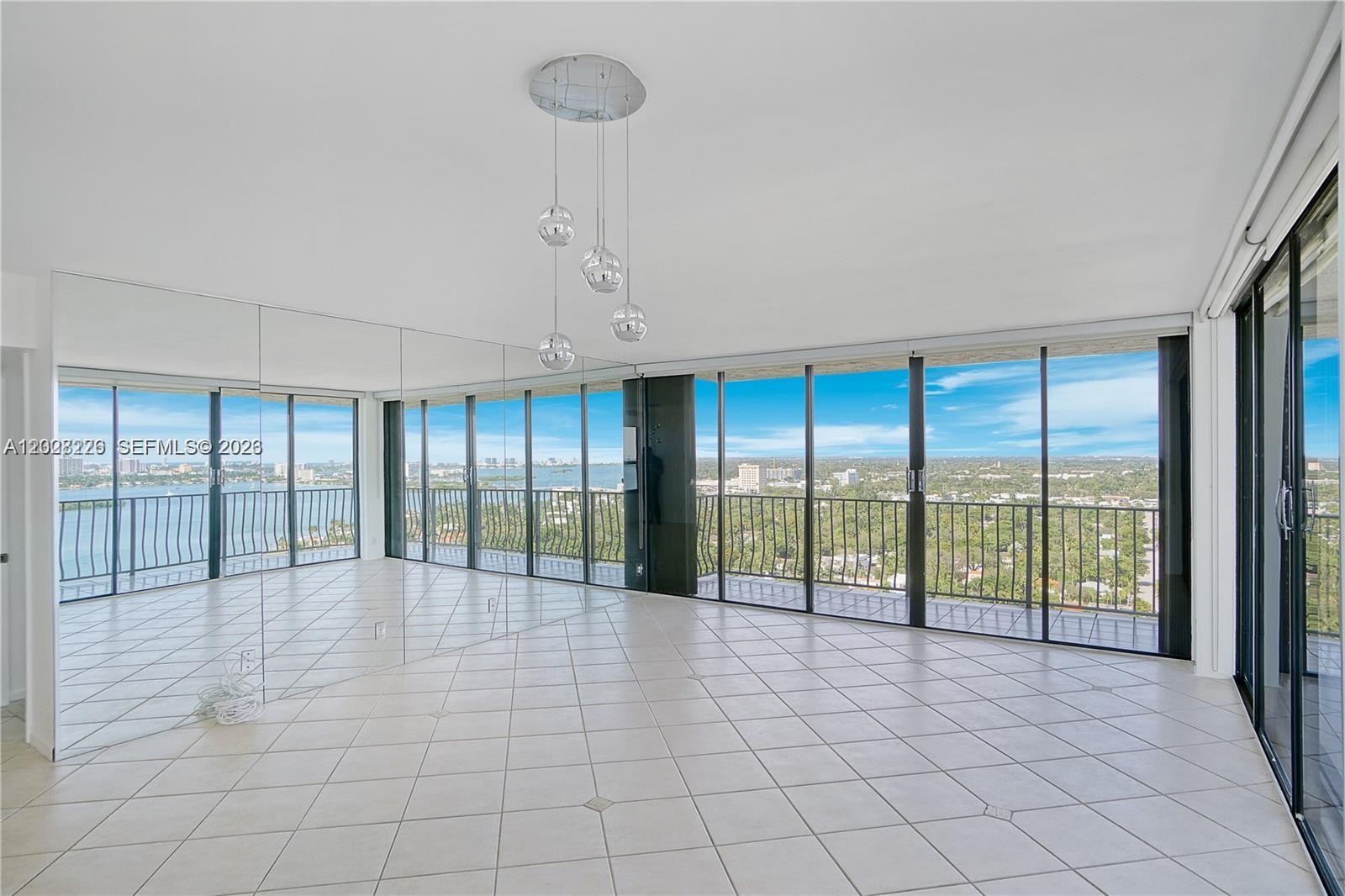 An empty room features large mirrored walls, white tile flooring, and floor-to-ceiling windows with views — 780 NE 69th St # 2302, Miami FL 33138, MIAMI, FL