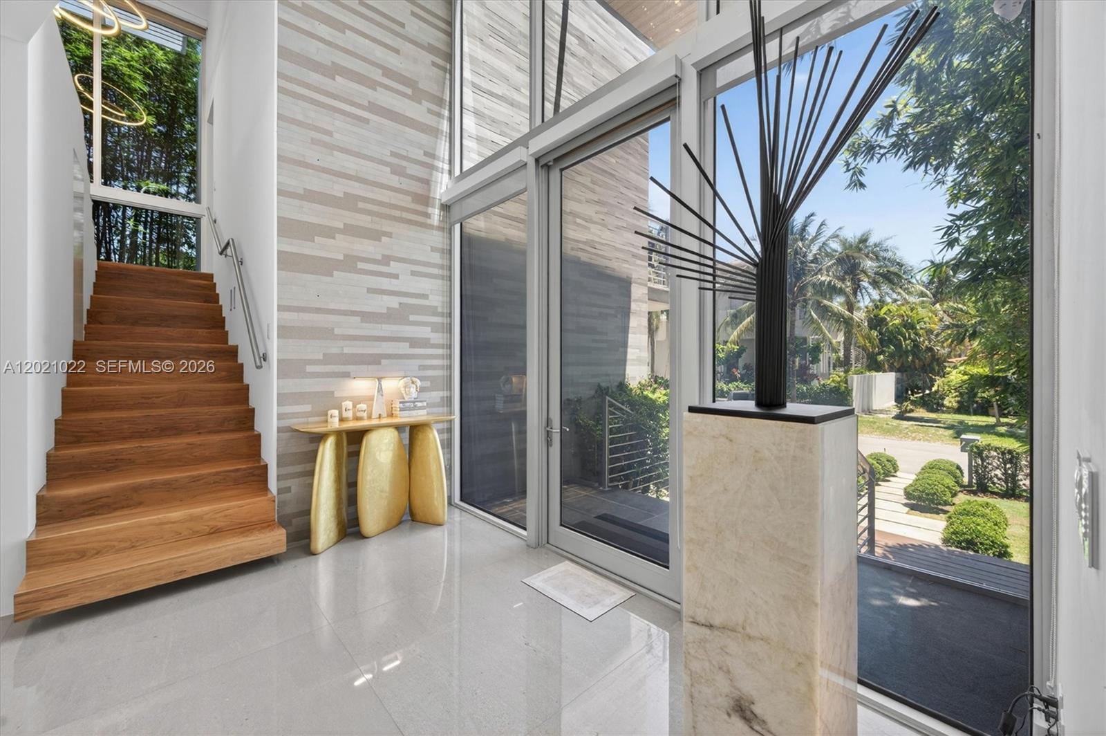 A modern entryway features a wooden staircase, a gold console table, and floor-to-ceiling glass windows — 625 Curtiswood Dr, Key Biscayne FL 33149, KEY BISCAYNE, FL