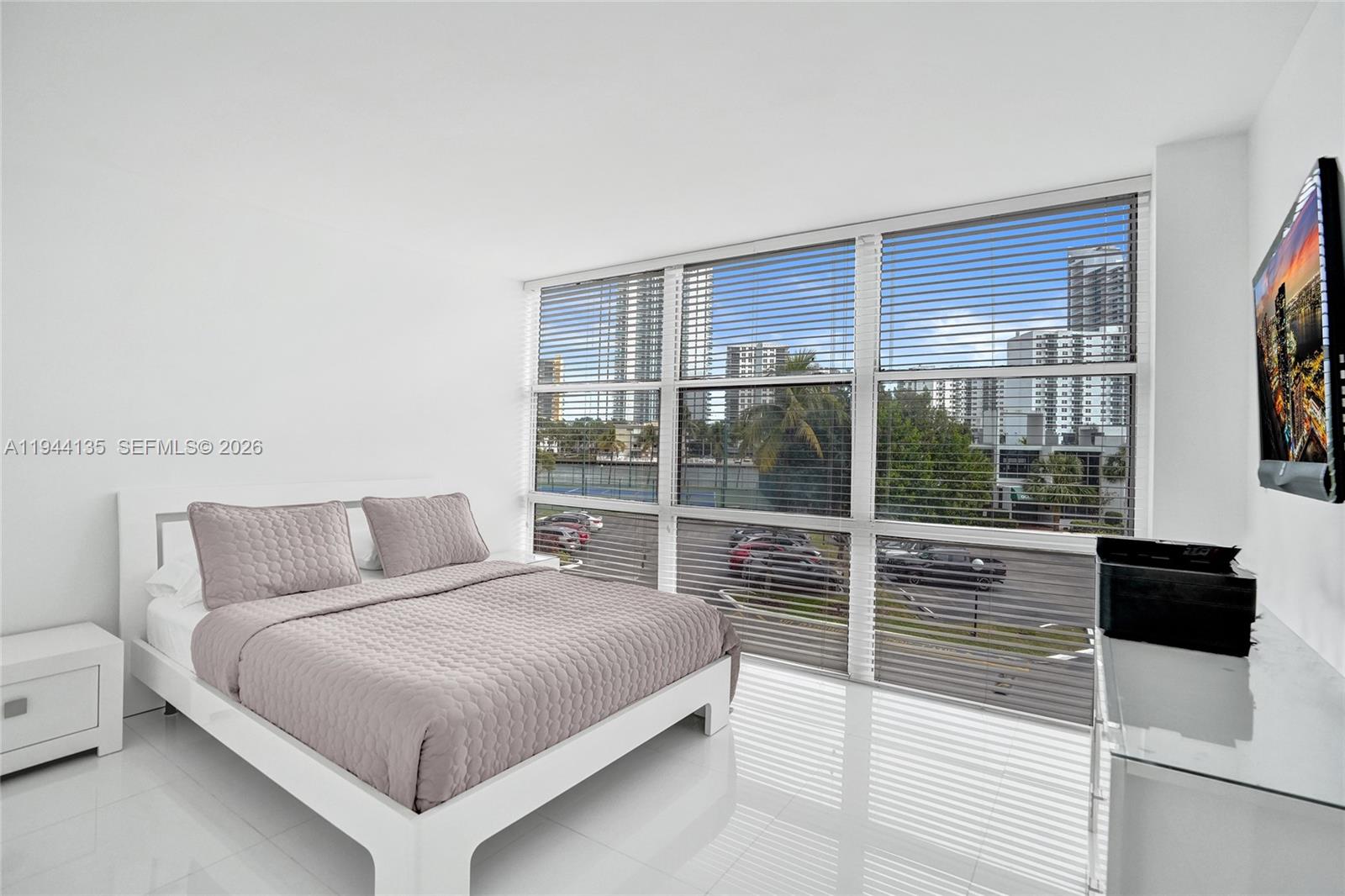 A modern bedroom features a white bed, tiled floors, and large windows overlooking a city — 1000 Parkview Dr # 330, Hallandale Beach FL 33009, HALLANDALE BEACH, FL
