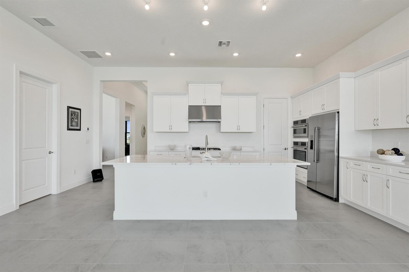 A modern white kitchen features a large center island, stainless steel appliances, and gray tile flooring — 5183 Rivo Alto Circle, Westlake FL 33470, WESTLAKE, FL