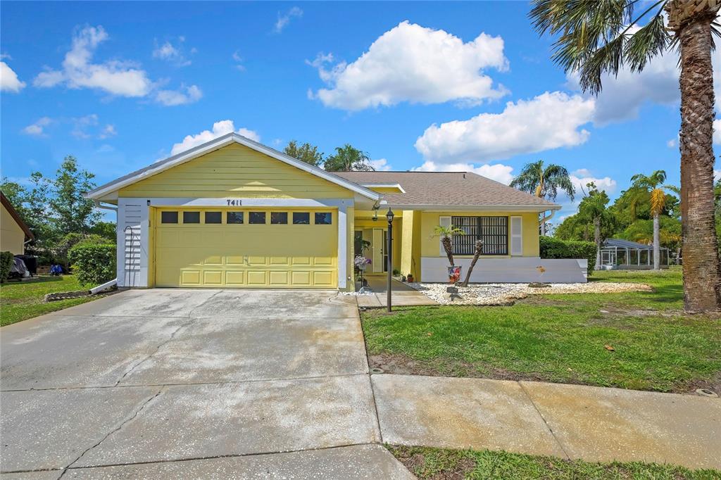 A yellow single-story house features a two-car garage, a concrete driveway, and a palm tree — 7411 BALTUSROL DRIVE, NEW PORT RICHEY, FL