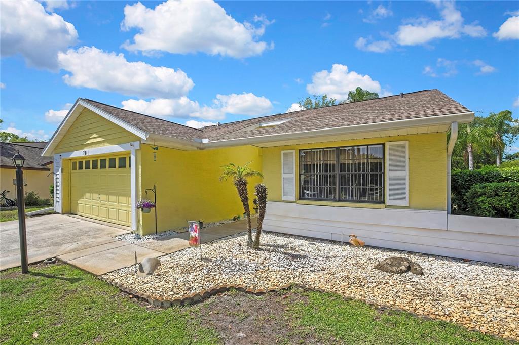 A yellow single-story house features a two-car garage and a rock-covered front yard — 7411 BALTUSROL DRIVE, NEW PORT RICHEY, FL