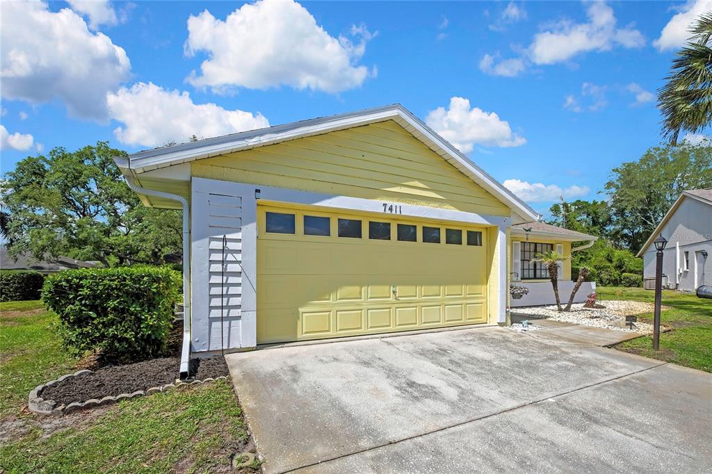 A yellow house with a two-car garage and a concrete driveway under a blue sky — 7411 BALTUSROL DRIVE, NEW PORT RICHEY, FL