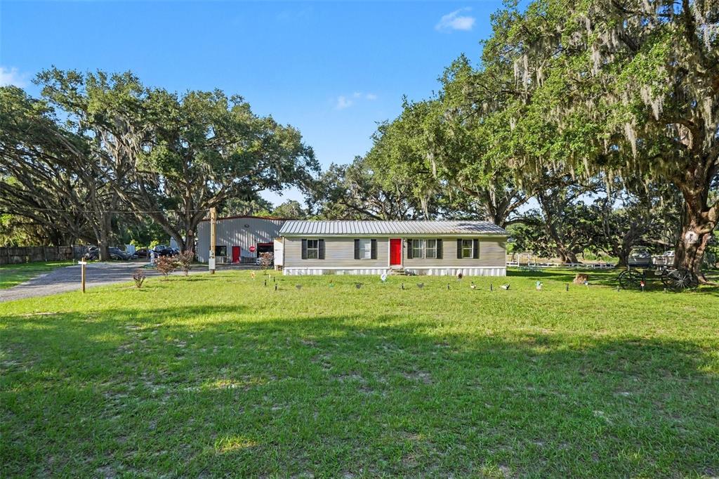 A single-story house with a red door and an adjacent metal workshop under large trees — 12709 CHIHUAHUA COURT, THONOTOSASSA, FL