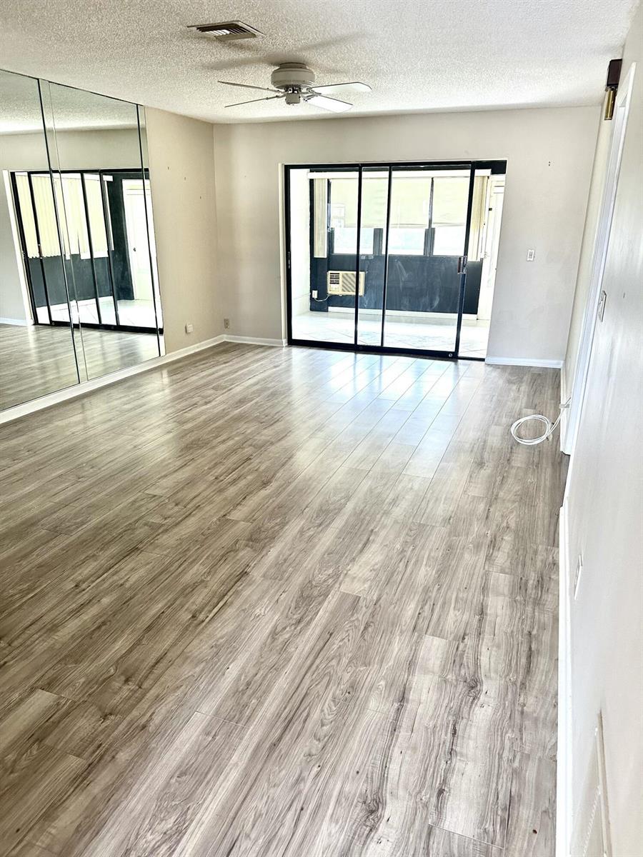 An empty living room features light wood-look flooring, a mirrored wall, and sliding glass doors — 6792 Moonlit Drive, Delray Beach FL 33446, DELRAY BEACH, FL
