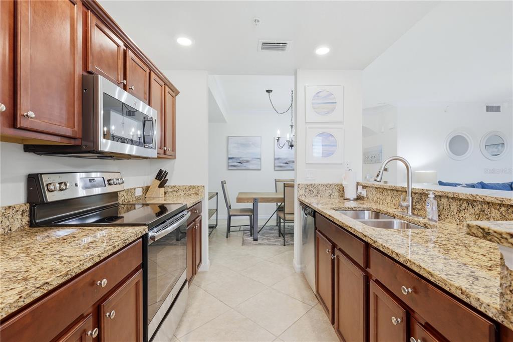 A kitchen with brown cabinets, granite countertops, and stainless steel appliances leads to a dining area — 16804 VARDON TERRACE UNIT 103, BRADENTON, FL