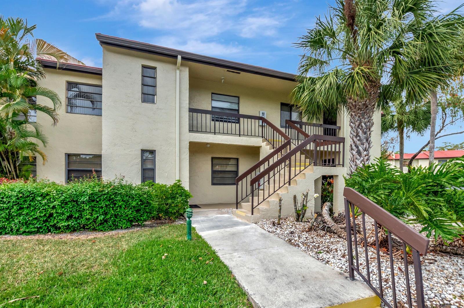A two-story apartment building with a concrete walkway, exterior stairs, and tropical landscaping — 21214 Lago Circle # 8f, Boca Raton FL 33433, BOCA RATON, FL