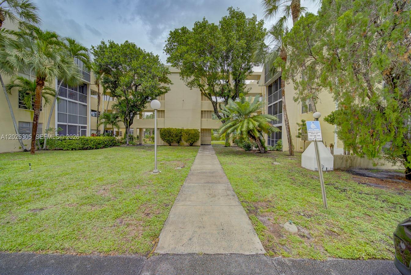A concrete walkway leads to the entrance of a multi-story apartment building with landscaping — 942 NE 199th St # 102, Miami FL 33179, MIAMI, FL