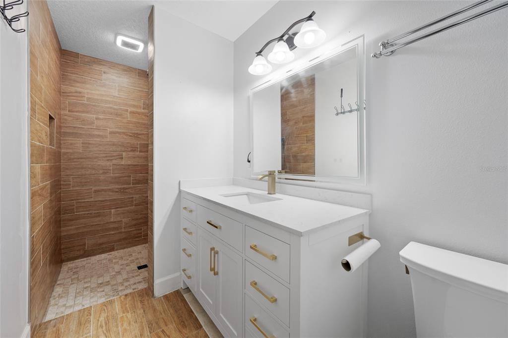 A bathroom features a white vanity, a walk-in shower with wood-look tile, and gold hardware — 4115 55TH AVENUE DRIVE E, BRADENTON, FL