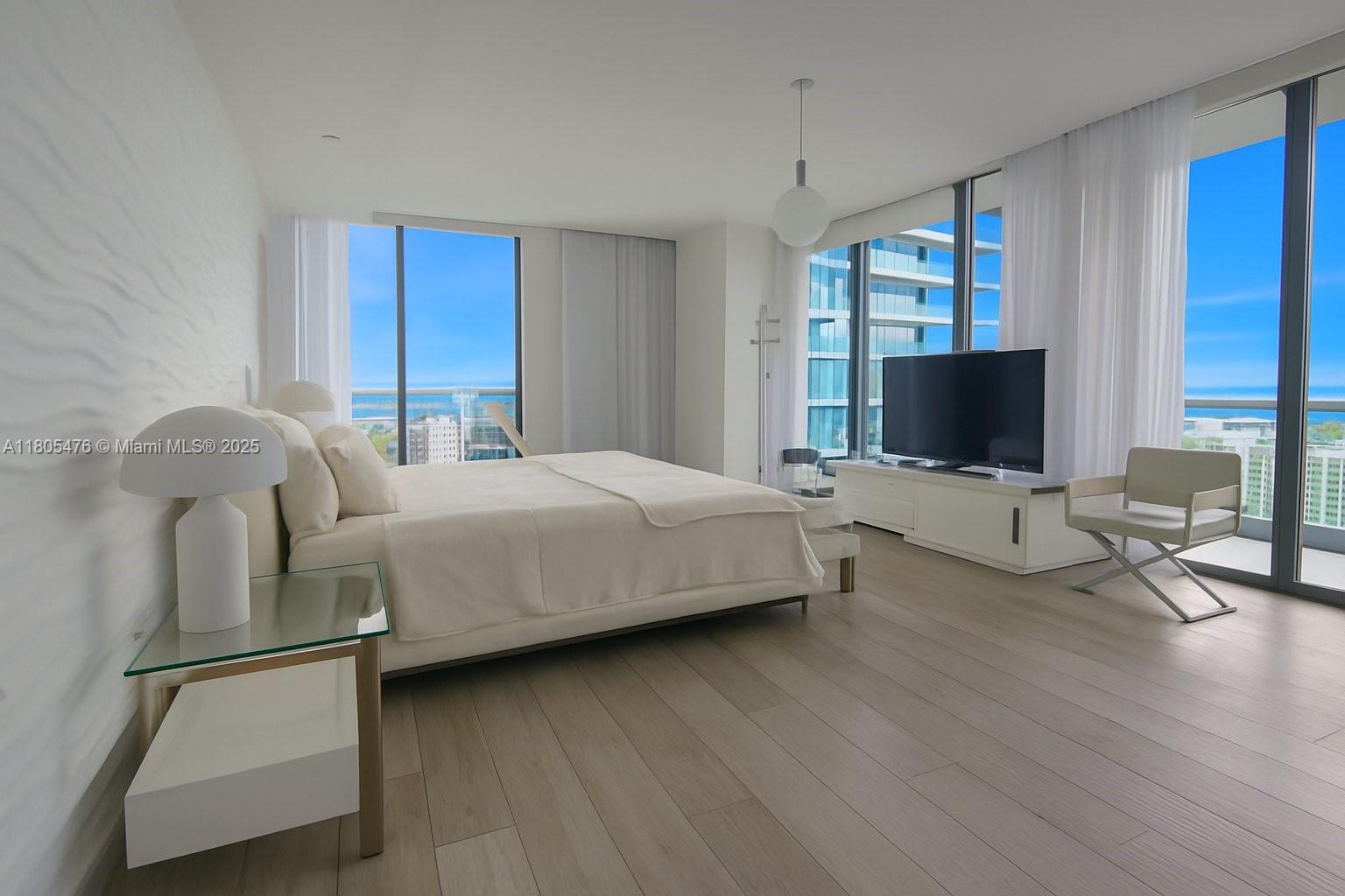A modern bedroom features light wood flooring, a white bed, and floor-to-ceiling windows with ocean views — 480 NE 31 St # 2607, Miami FL 33137, MIAMI, FL