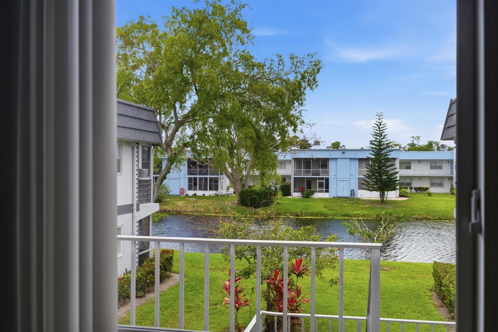 A balcony overlooks a small pond, green lawn, and blue apartment buildings with mature trees — 376 Saxony H, Delray Beach FL 33446, DELRAY BEACH, FL