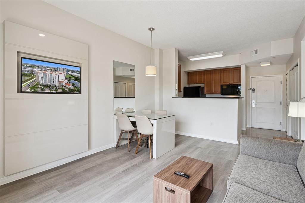 A living and dining area with light wood floors, a wall-mounted television, and kitchen access — 7395 UNIVERSAL BOULEVARD UNIT 403, ORLANDO, FL