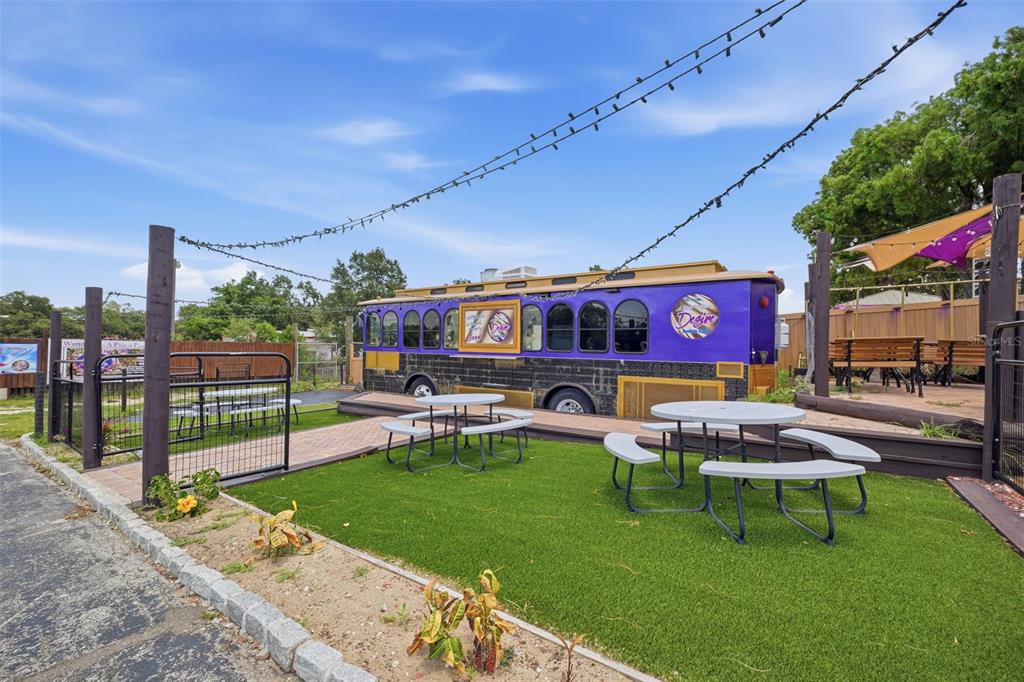 An outdoor patio features a purple bus, picnic tables, and string lights on artificial turf — 8744 GALL BLVD, ZEPHYRHILLS, FL