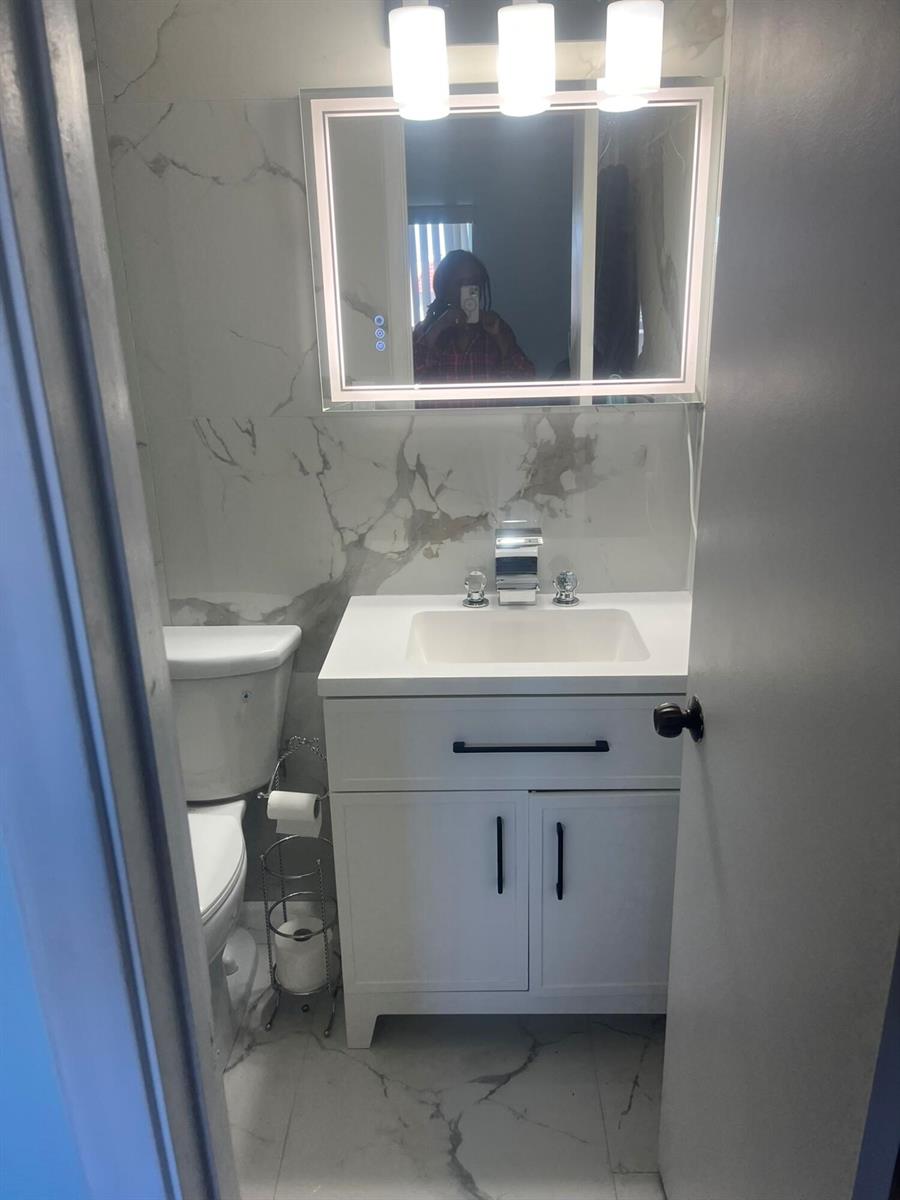 A small bathroom features a white vanity, marble-patterned wall tiles, and a lighted mirror — 3531 Inverrary Drive # 308, Lauderhill FL 33319, LAUDERHILL, FL