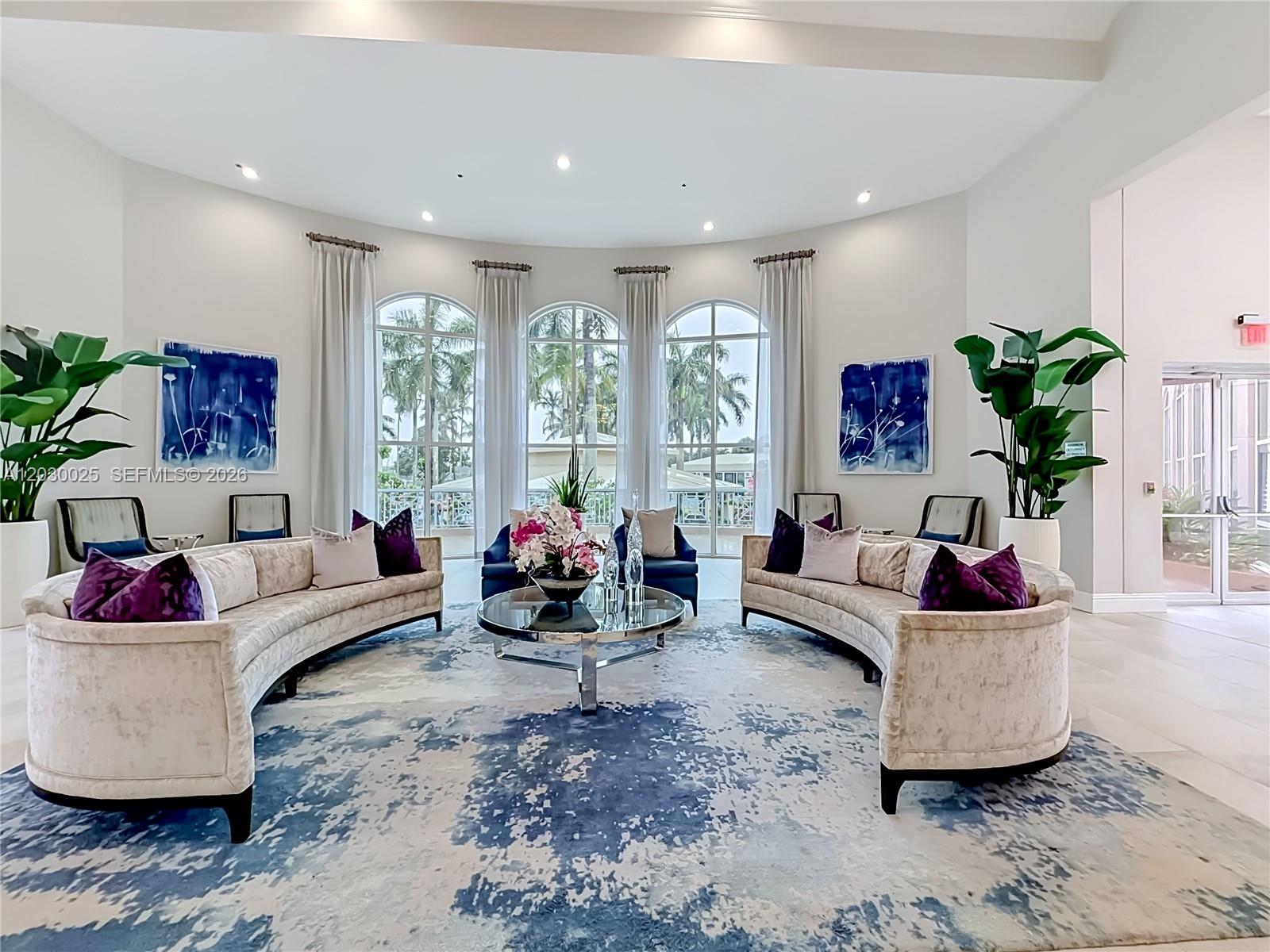 A bright, circular lounge features two curved sofas, a glass coffee table, and arched windows — 5591 Royal Lake Cir, Boynton Beach FL 33437, BOYNTON BEACH, FL