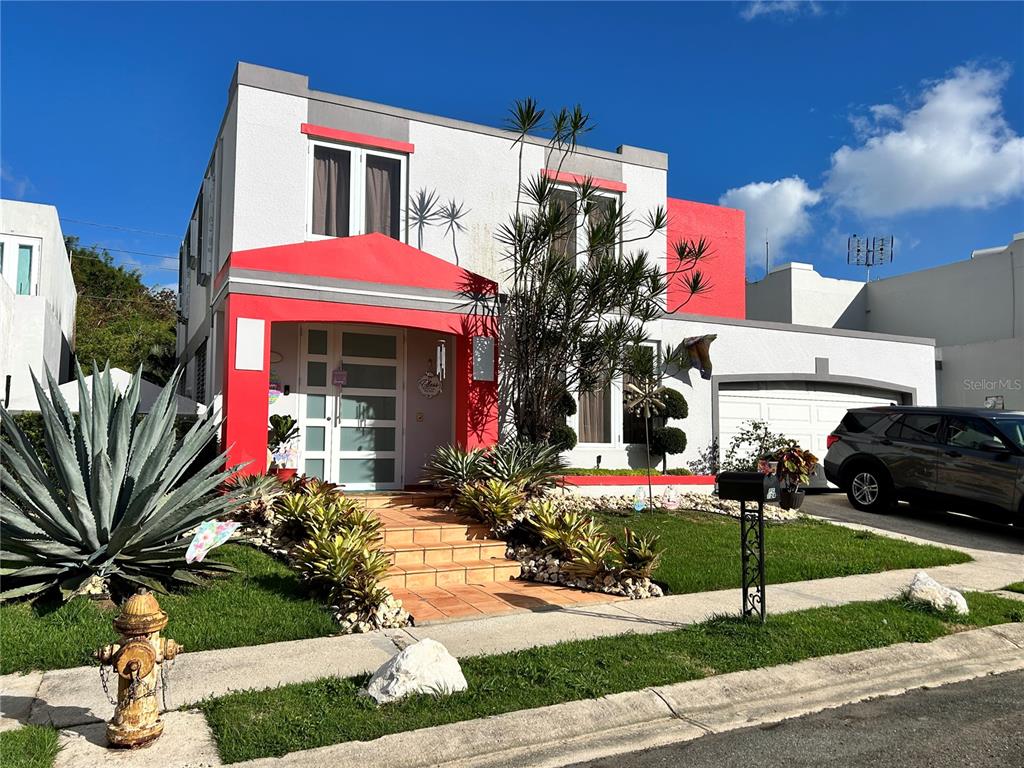 A two-story house features a red-accented entryway, a front lawn, and a parked SUV — PG-124 VIA ARCOIRIS, TRUJILLO ALTO, PR