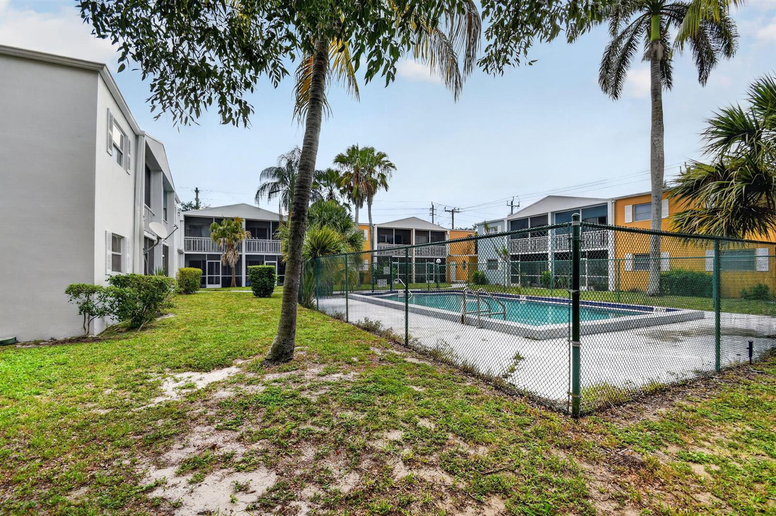 A fenced swimming pool sits on a lawn between multi-story apartment buildings with palm trees — 1505 Crescent Circle # B24, Lake Park FL 33403, LAKE PARK, FL