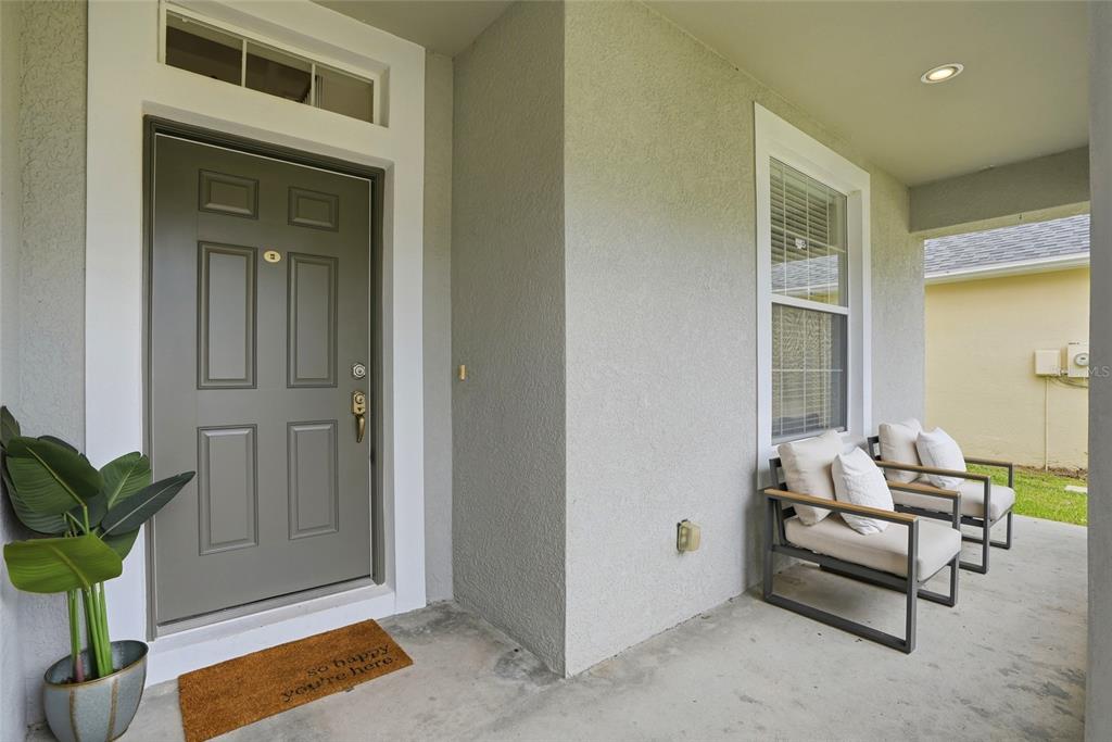 A covered front porch features a gray door, a welcome mat, and two outdoor chairs — 3203 SUNWATCH DRIVE, WESLEY CHAPEL, FL