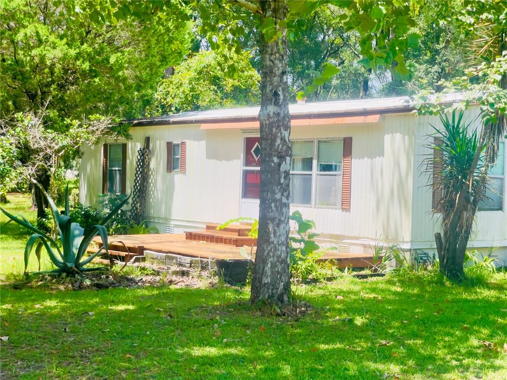 A single-story mobile home with a wooden deck sits in a grassy, tree-filled yard — 459 MARSHBURN DR, BRONSON, FL