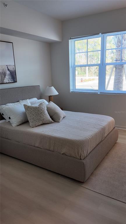 A bedroom features a gray upholstered bed, light wood floors, and a window with blinds — 5587 MILLENIA PARK BLVD UNIT 218, ORLANDO, FL