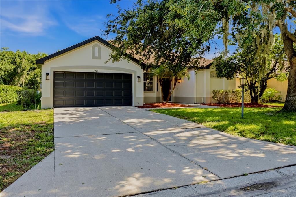 A single-story house with a dark garage door and a concrete driveway under a tree — 1710 BROOKSTONE WAY, PLANT CITY, FL
