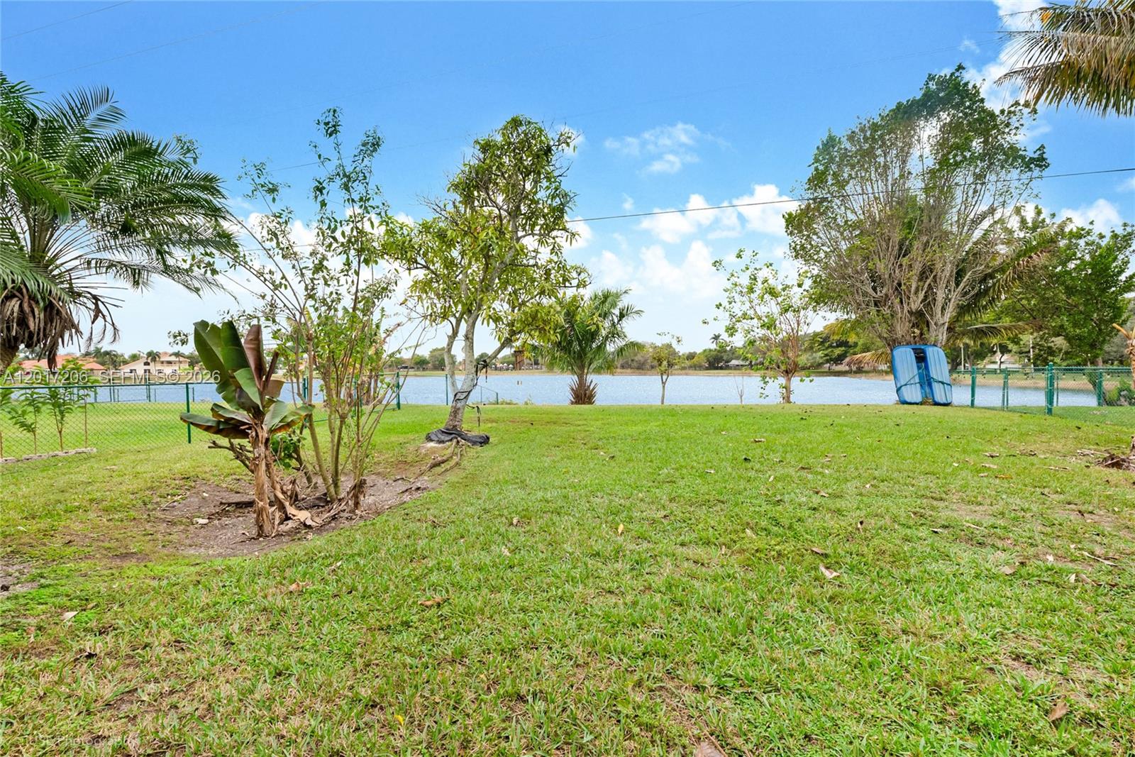 A grassy backyard features trees, a fence, and a view of a lake — 1402 SE 16th Ave, Homestead FL 33035, HOMESTEAD, FL