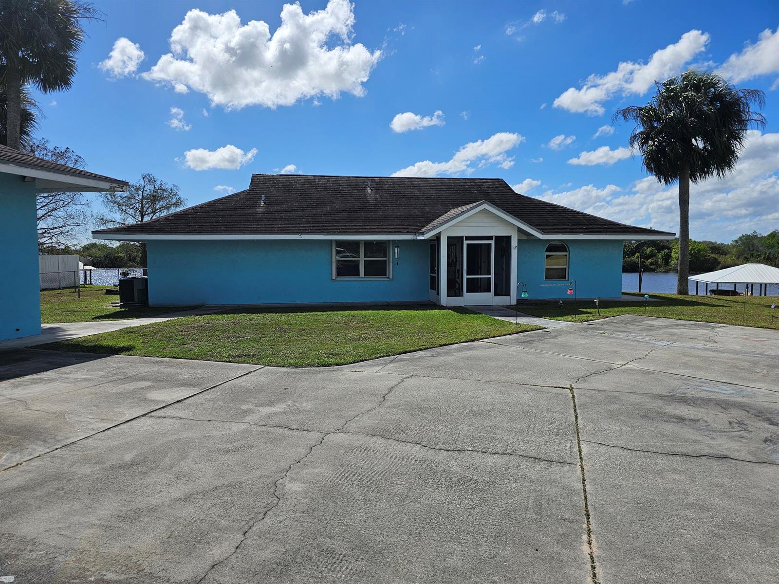 A blue, single-story house with a dark shingled roof sits beside a body of water — 13578 SW 144th Parkway, Okeechobee FL 34974, OKEECHOBEE, FL