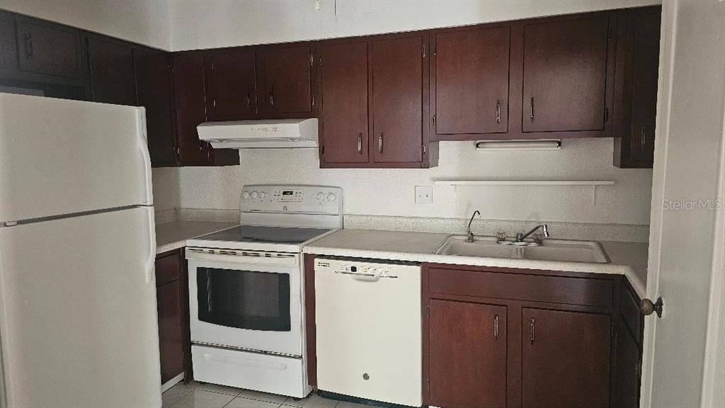 A kitchen features dark wood cabinets, white appliances, and a white countertop with a sink — 10365 PARADISE BOULEVARD UNIT 10, TREASURE ISLAND, FL