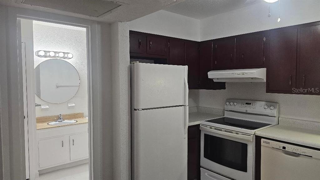 A kitchen with dark wood cabinets and white appliances sits adjacent to a small bathroom — 10365 PARADISE BOULEVARD UNIT 10, TREASURE ISLAND, FL