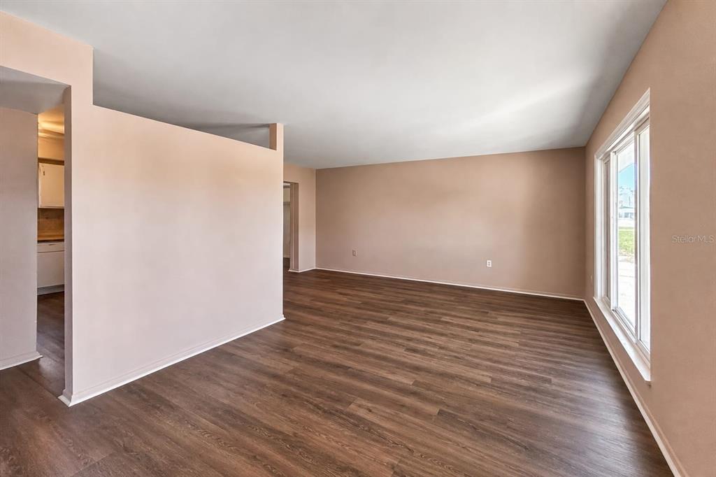 An empty living room features light-colored walls, dark wood-look flooring, and a large window — 1515 ARROWHEAD DRIVE, SUN CITY CENTER, FL
