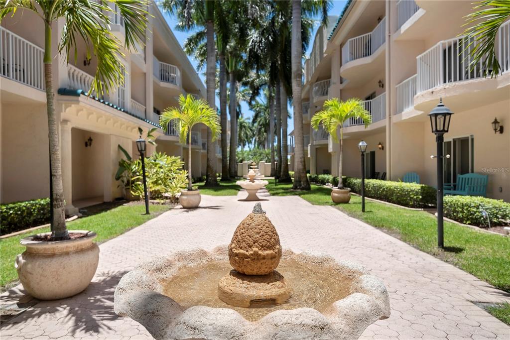 A stone fountain sits on a paved walkway between two multi-story apartment buildings with palms — 5963 MIDNIGHT PASS ROAD UNIT 332, SARASOTA, FL