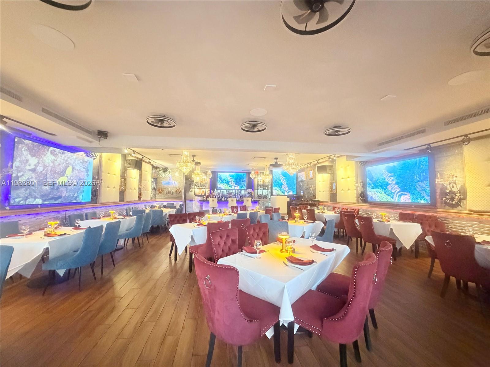 A restaurant dining room features white-clothed tables, red and blue upholstered chairs, and large screens — SUNNY ISLES BEACH, FL