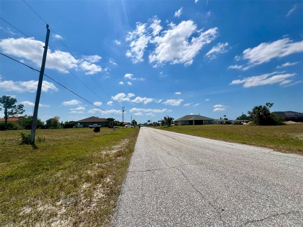 A paved road runs between two grassy vacant lots under a bright blue sky — 1031 NW 20TH PLACE, CAPE CORAL, FL