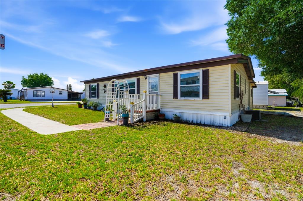 A light yellow manufactured home with brown shutters sits on a grassy lot — 3933 SARAH DRIVE, WESLEY CHAPEL, FL
